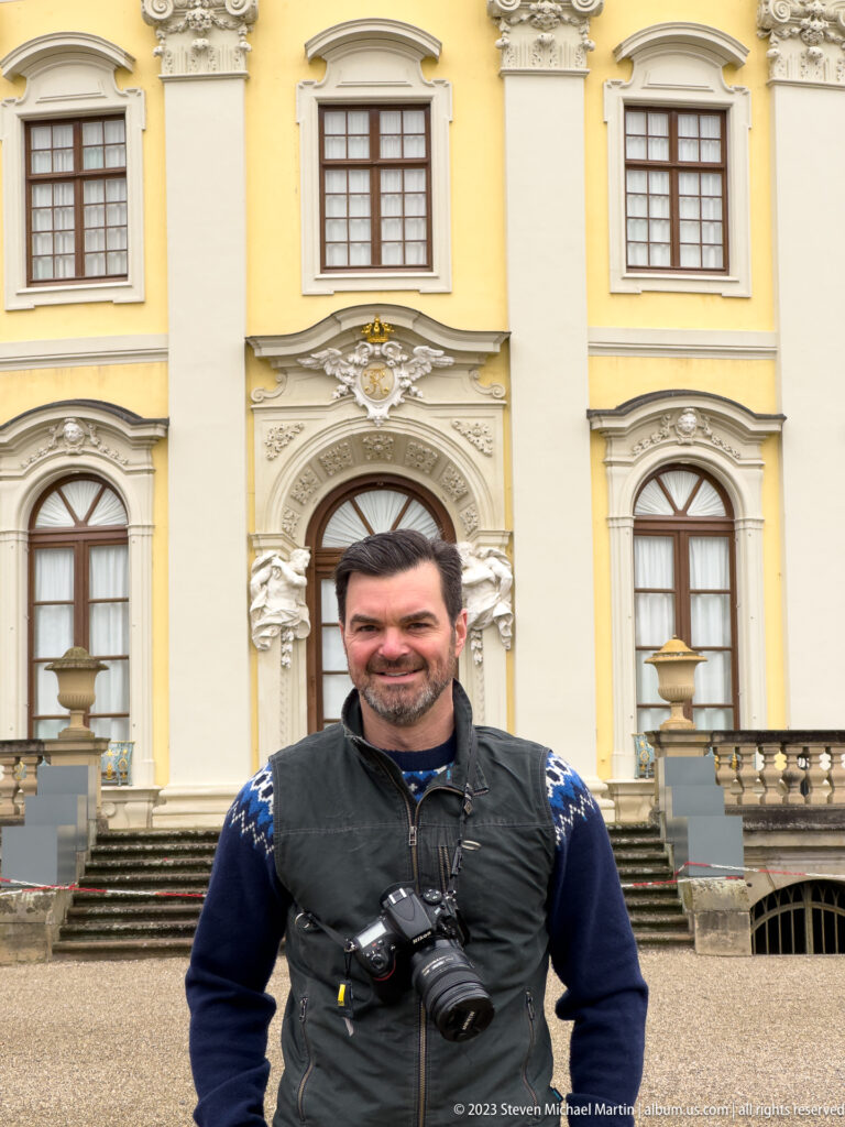 Visitor in front of a yellow palace entrance bay with carved stone ornament, arched windows, and a short staircase.
