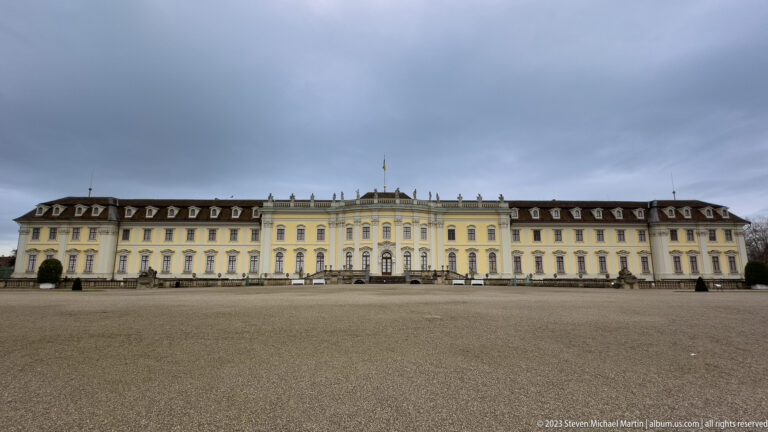 Long symmetrical yellow-and-white palace façade across a broad gravel forecourt beneath a gray sky.