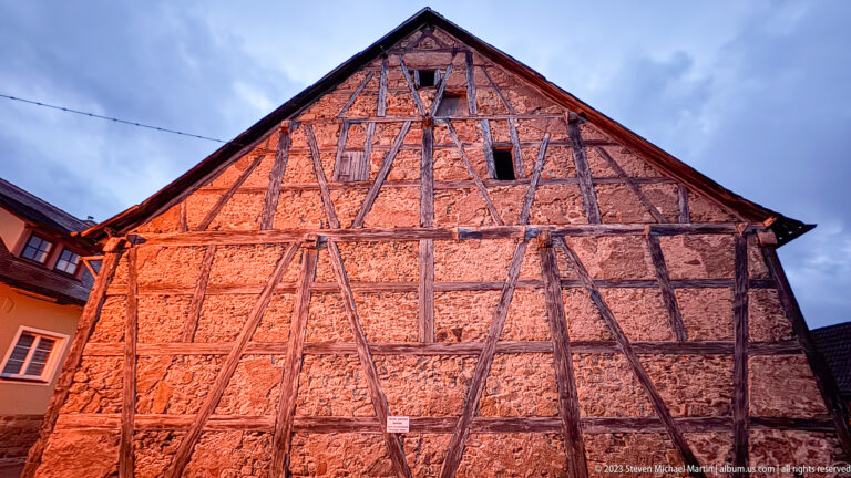 Steep half-timbered gable with reddish infill, irregular timber braces, small upper openings, and neighboring roofs under a blue-gray sky.