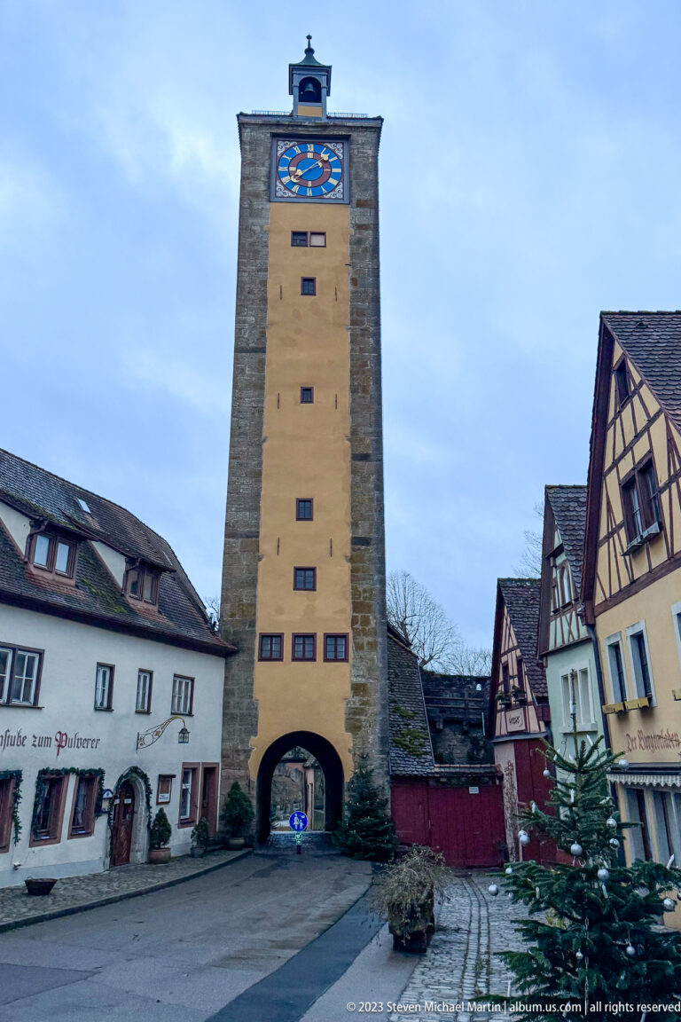 Tall rectangular stone gate tower with an arched passage and clock face, flanked by historic houses under an overcast sky.