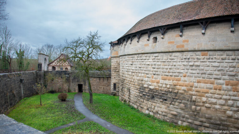 Curving high stone fortification wall beside a grassy enclosed space and branching path, with a smaller tower and gate beyond.