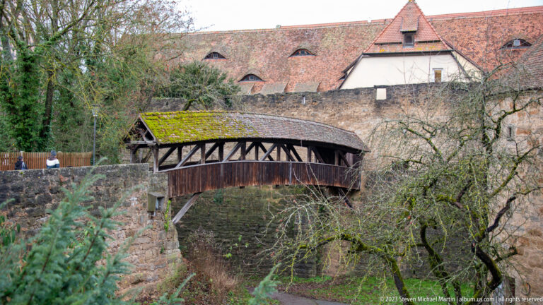 Small covered timber footbridge with diagonal braces and mossy roof spanning a lower passage beside stone fortifications.