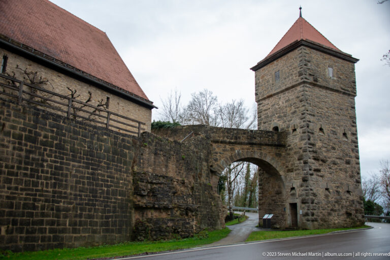 Square stone gate tower with rounded road arch, small openings, red-tiled roof, and adjoining fortification wall.