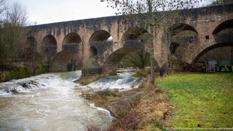Doppelbrücke over rushing white water in the Tauber River, with massive piers, broad lower arches, and smaller upper arches.