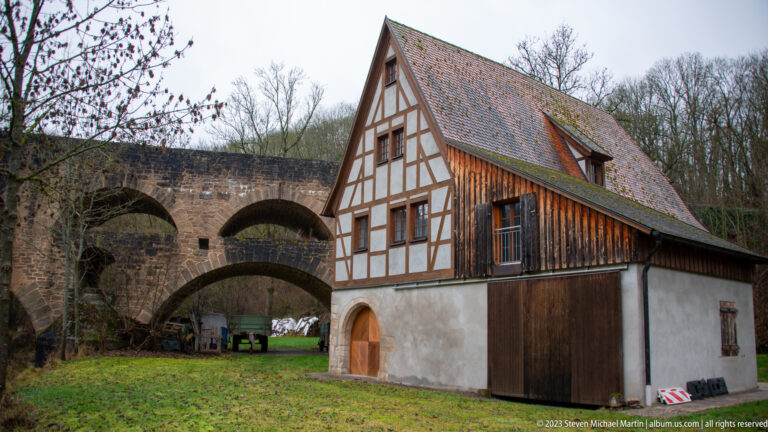 Steep-gabled half-timbered riverside building with wooden side cladding, standing beside the stone arches of the Doppelbrücke.