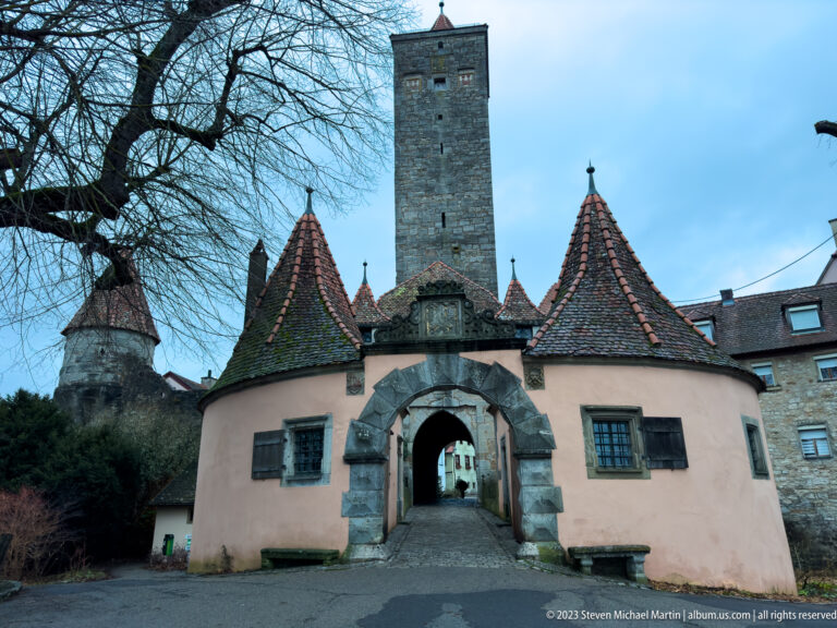 Low stuccoed gatehouse with steep tiled roofs and small turrets beneath a tall square stone tower and arched entrance.
