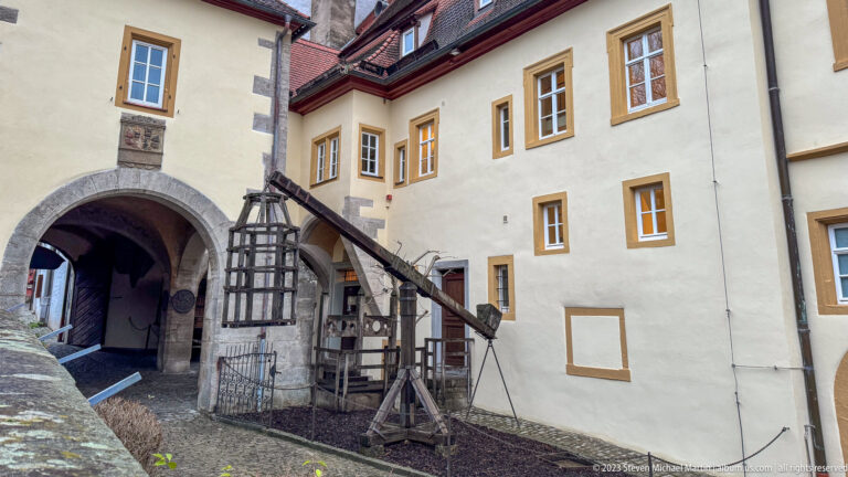 Large historic timber hoist with beams, braces, chain, and winding mechanism beside an arched passage and plastered building.