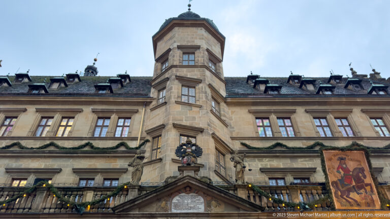 Close low-angle view of the Rathaus Renaissance façade with projecting polygonal bay, stone ornament, heraldic emblem, and festive garlands.