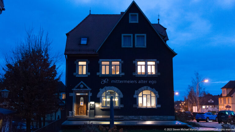 Dark gabled building marked “mittermeiers alter ego,” with warm illuminated windows, parked cars, and street lighting at blue hour.