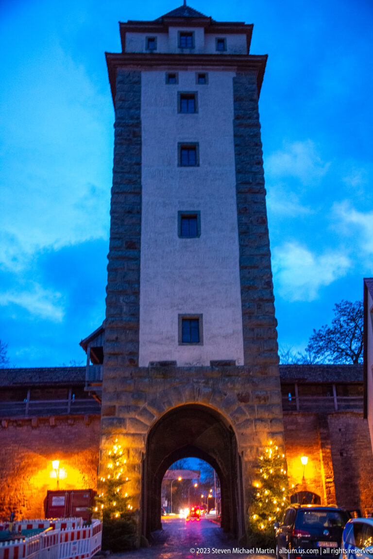 Large fortified gate tower with central arch, stone base, pale upper shaft, broad roof, Christmas trees, and warm evening lights.