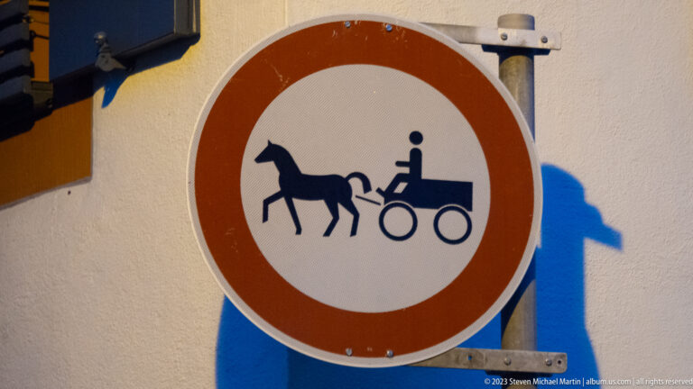 Round red-bordered traffic sign with a black silhouette of a horse pulling a carriage against a pale wall.