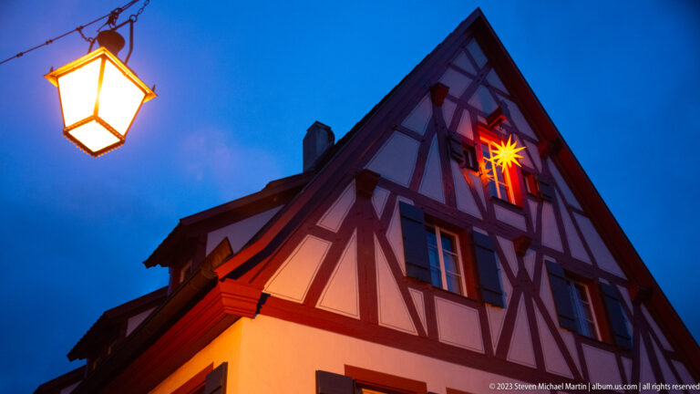 Steep half-timbered gable lit warmly at dusk, with a glowing star decoration in an upper window and a streetlamp at left.