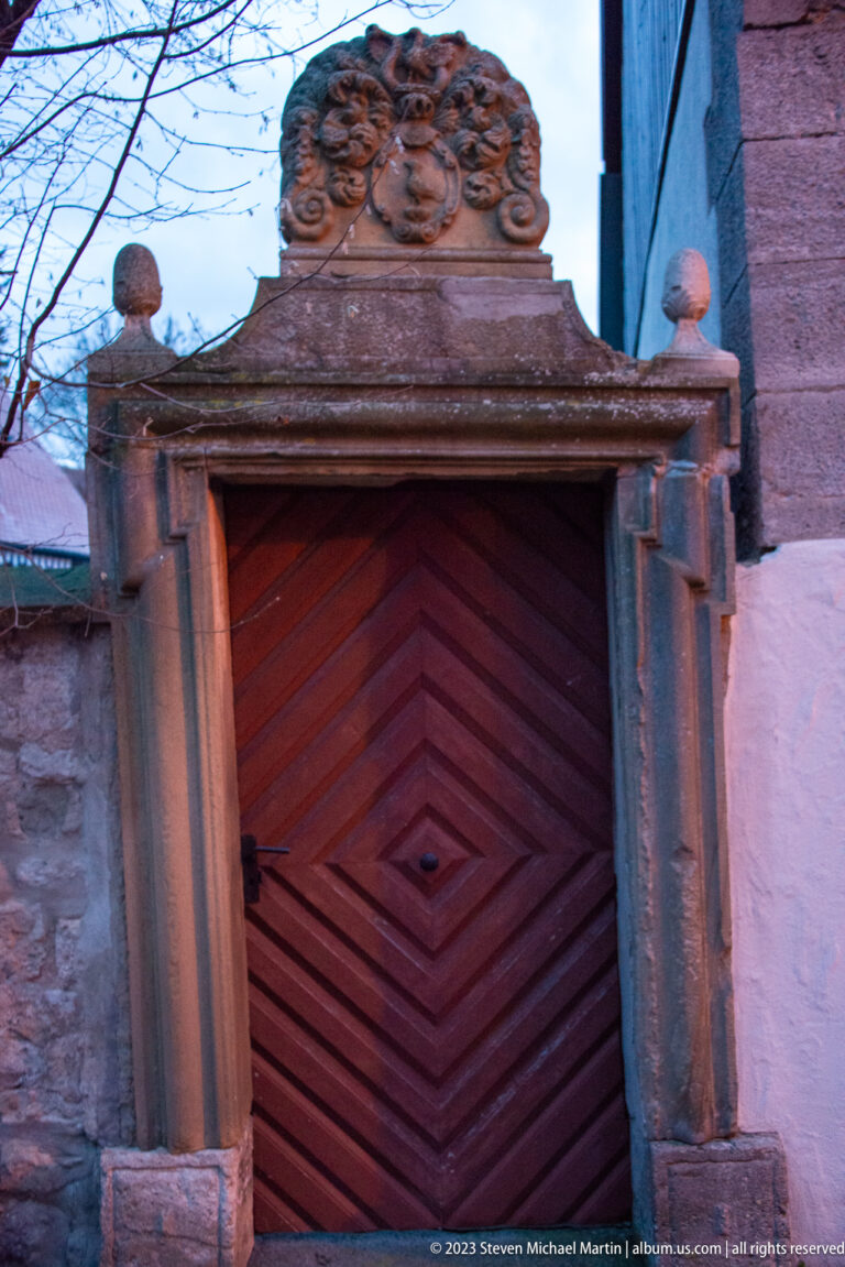 Reddish chevron-patterned timber door within a carved stone portal topped by an ornamental relief panel.