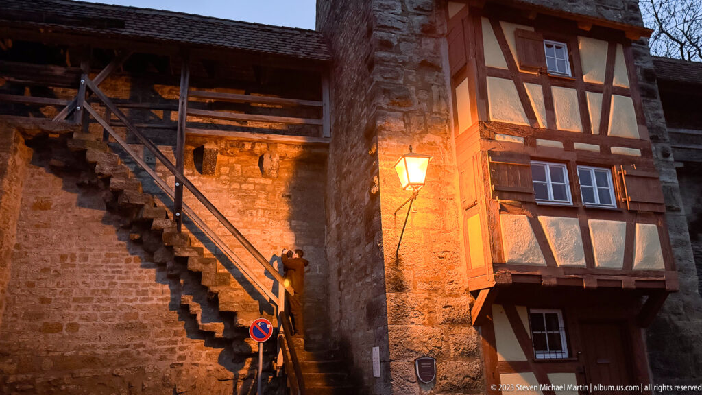 Wooden stair beside a high stone fortification wall and projecting half-timbered house, illuminated by a warm streetlamp at dusk.