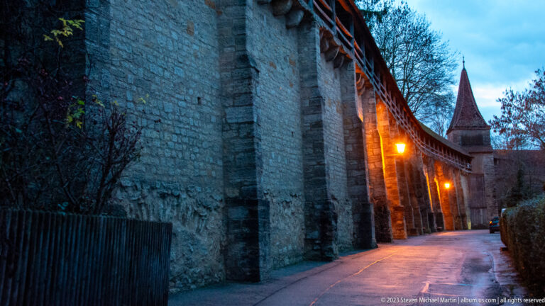Narrow lane beside a high stone city wall and covered wall walk, lit by warm lamps with a pointed-roof tower ahead.