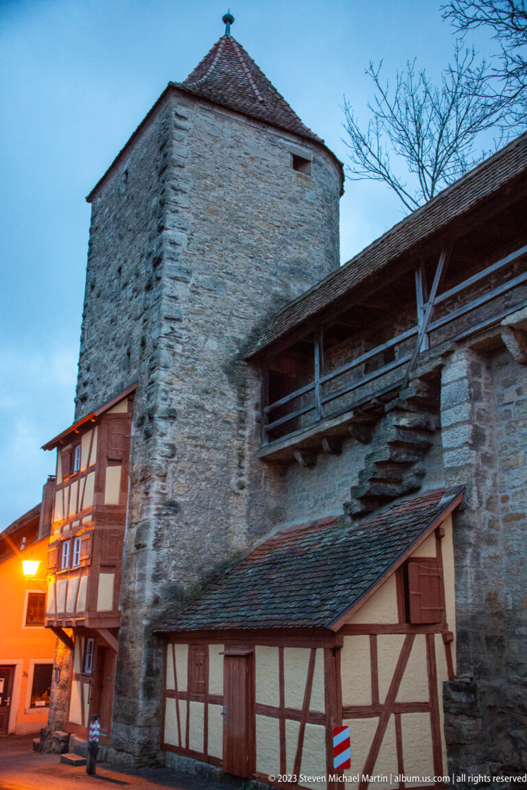 Broad stone defensive tower with covered wall walk and small half-timbered buildings at its base, seen at dusk.