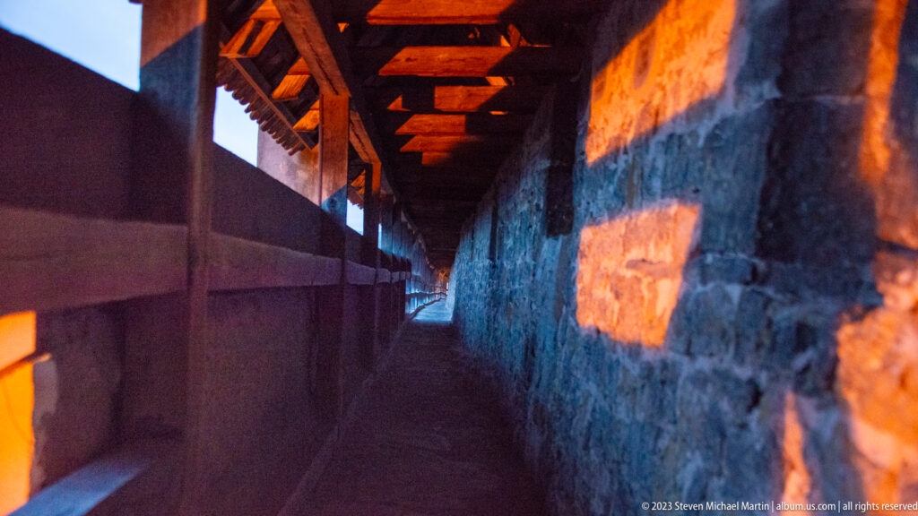 Interior of a covered city-wall walk with timber parapet, repeating roof beams, rough stone wall, and warm light patches.