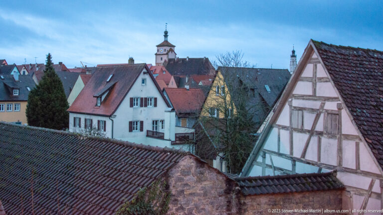 Elevated view across red tile roofs, pale houses, a half-timbered gable, and a distant tower in Rothenburg's old town.