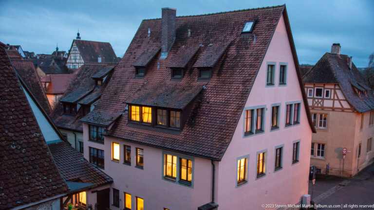 Pale gabled house with steep red-tile roof, dormers, chimney, and glowing windows, surrounded by historic roofs at dusk.