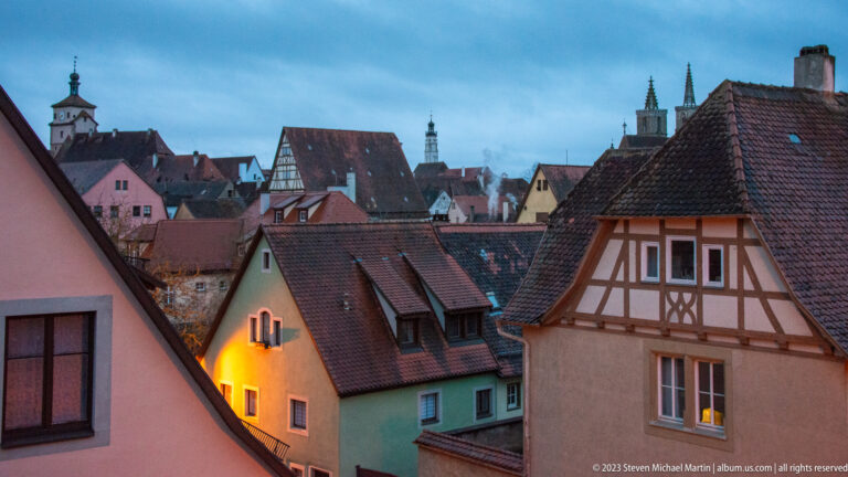 Layered pastel houses and steep tiled roofs with a half-timbered gable, distant towers, and warm lights at dusk.