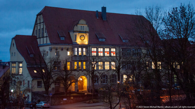 Large historic gabled building with red-tile roof, arched entrance, rows of windows, and an illuminated circular clock or medallion.