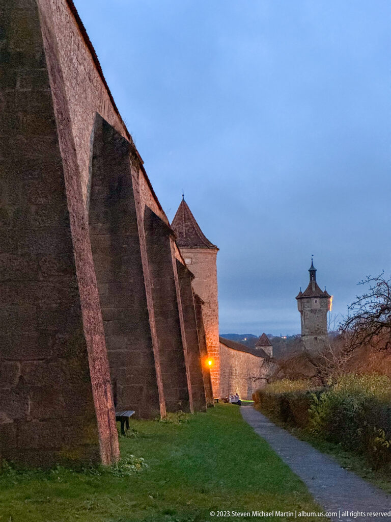 High stone city wall with angled supports, roofed defensive towers, grassy outer verge, and a path following the fortifications.