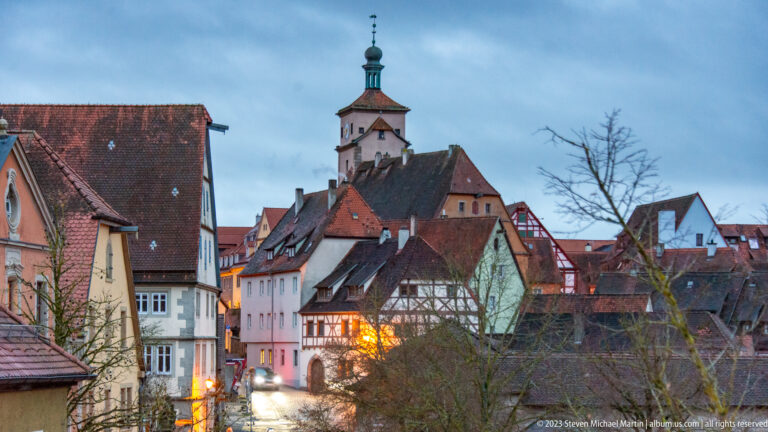 Narrow old-town street lined with steep-roofed and half-timbered houses, warm lights, and a prominent tower above the rooftops.