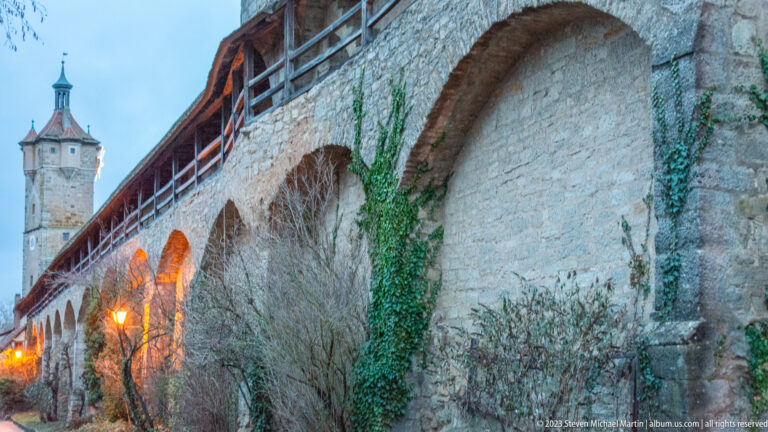 Long stone city wall with broad arches, ivy, covered timber wall walk, warm lamps, and a defensive tower beyond.