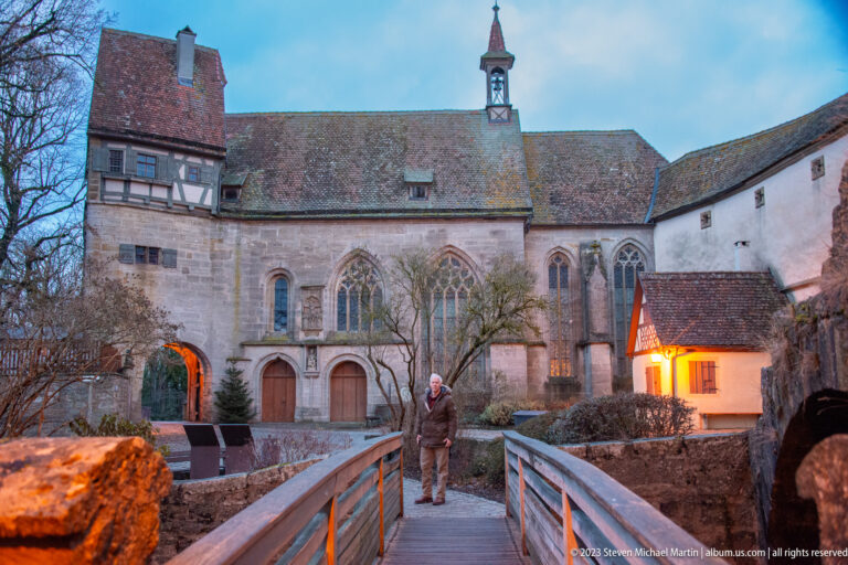 Gothic stone church with traceried windows, attached half-timbered section, arched passage, footbridge, and one visitor at dusk.