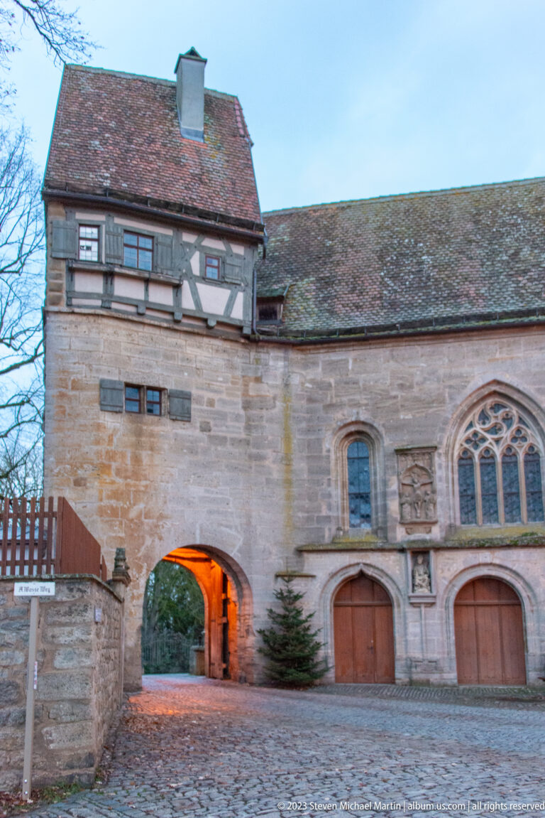 Stone church side wall with pointed traceried windows, broad wooden doors, arched passage, and half-timbered upper story.