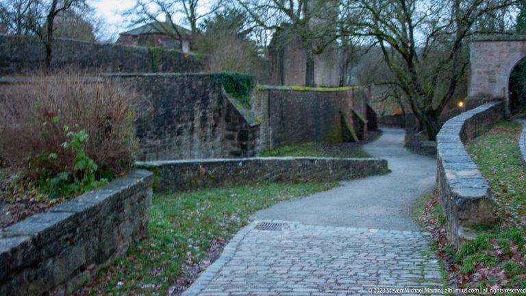 Curving paved path descending between low stone walls, grass, bare trees, and higher fortification masonry.