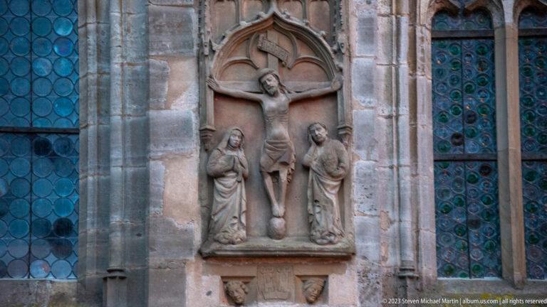 Stone Crucifixion relief with Christ and two standing figures beneath a pointed canopy between tall leaded-glass windows.