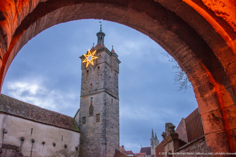 Square stone tower framed by a warm-lit arch, with a glowing Christmas star near its upper stage against the blue evening sky.