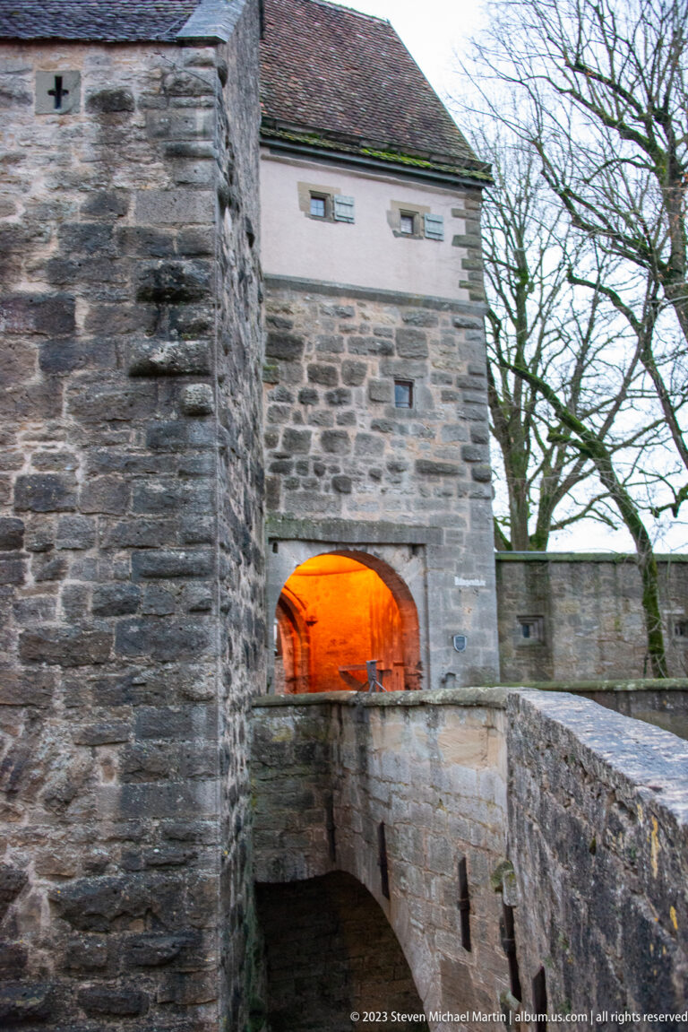 Stone gateway with a warmly illuminated arch reached by a narrow bridge between heavy fortification walls.