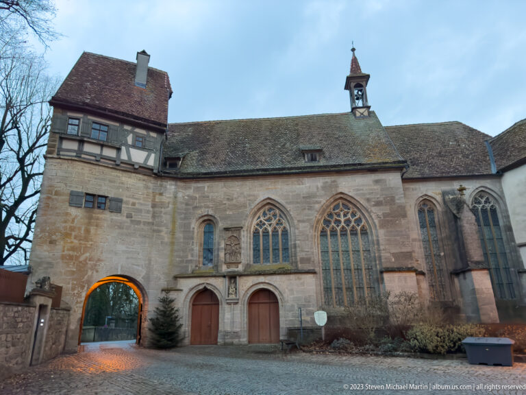 Gothic stone church with tall traceried windows, small bell turret, arched gateway, and adjoining half-timbered section at the city wall.