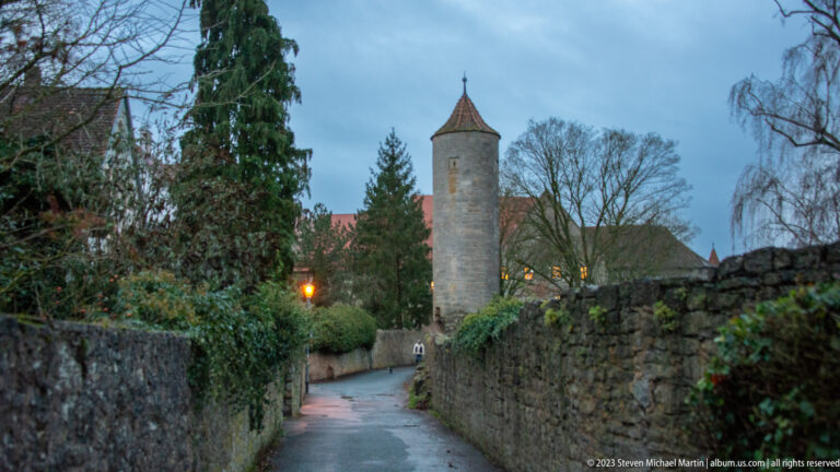 Narrow lane between stone walls leading toward a slender round tower with a conical roof and a glowing streetlamp.