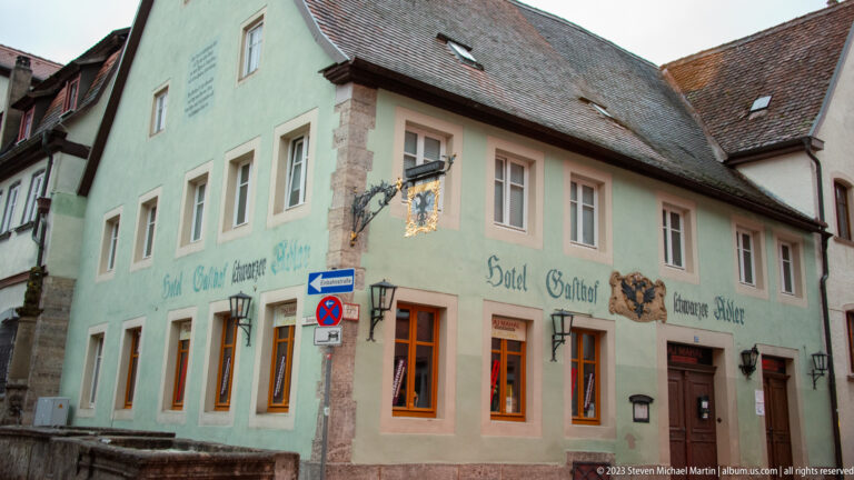Pale green corner hotel or inn with painted “Hotel Gasthof” lettering, hanging sign, tiled roof, and warm windows.