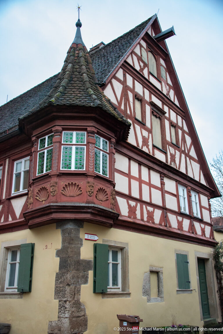 Decorated half-timbered corner house with red framing, projecting polygonal bay, green shutters, and a small conical roof.