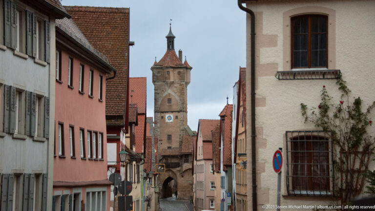 Narrow street lined with pastel houses leading to a tall stone gate tower with an arch, clock face, and steep roof.