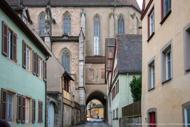 Narrow lane between pastel houses leading beneath an arch beside a tall Gothic church with pointed windows.