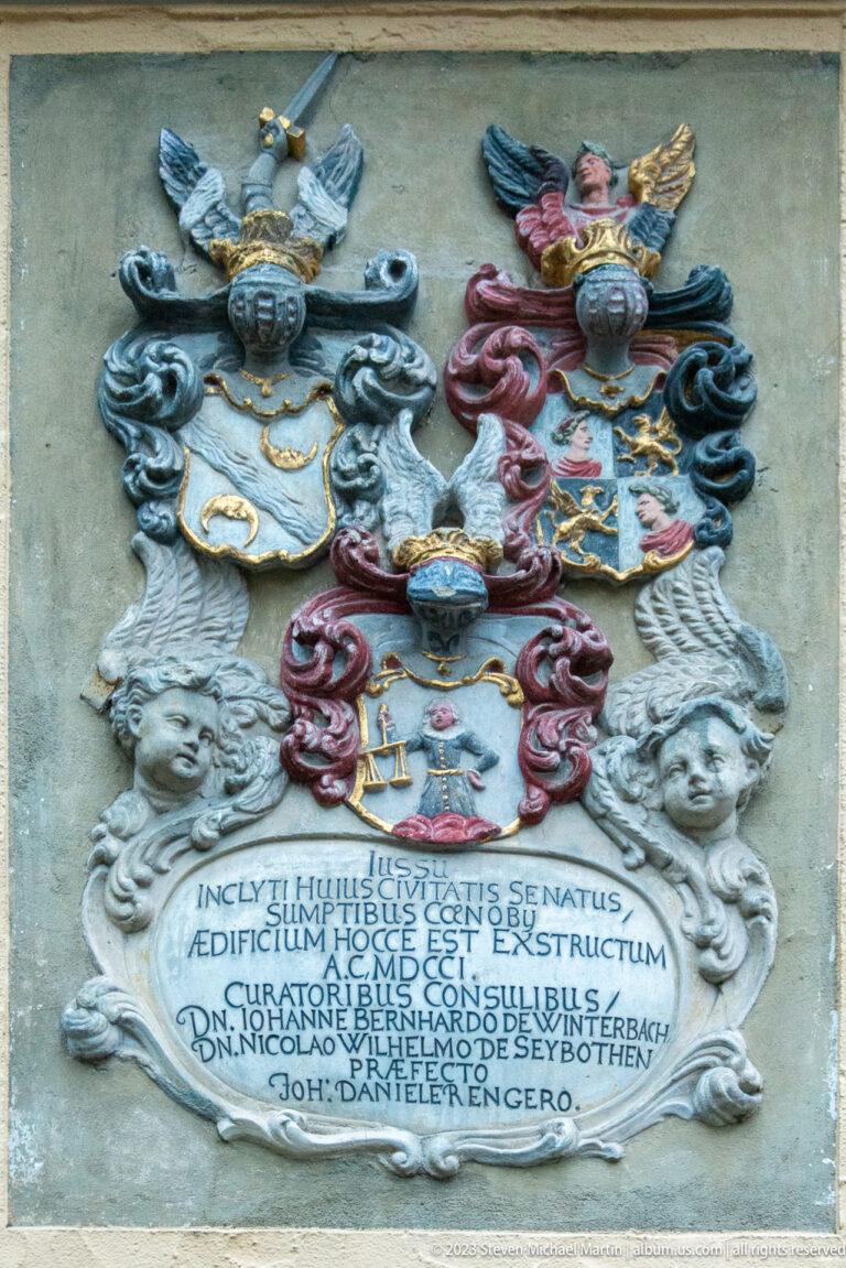 Ornate heraldic wall plaque with two colored coats of arms, helmets, scrolling mantling, faces, and a Latin inscription.
