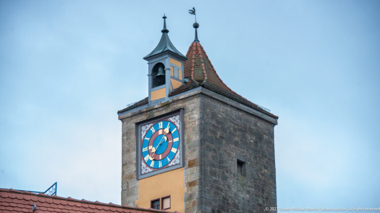 Close view of a colorful blue, red, white, and gold clock face on a square stone tower with two small roofed lanterns.