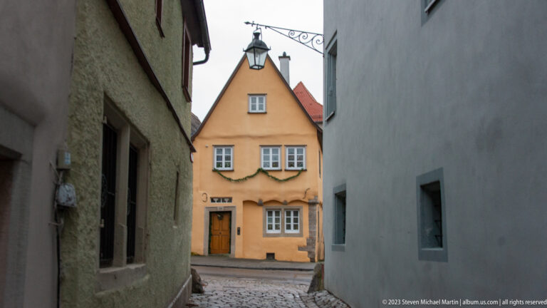 Narrow cobbled passage opening onto an ocher gabled house decorated with green garland and a hanging metal sign.
