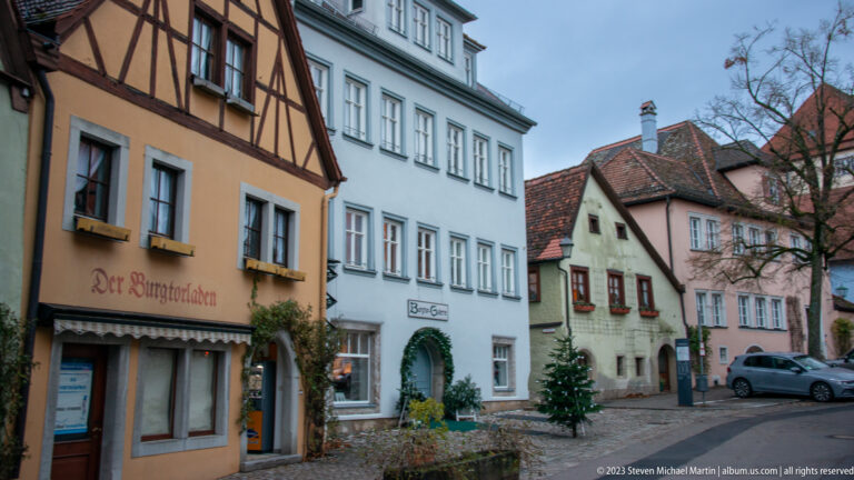 Small historic street corner with half-timbering, pastel façades, shop signs, Christmas trees, and a parked car.
