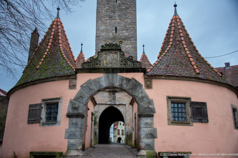 Pink gatehouse with a broad stone arch, twin round conical-roofed turrets, carved crest panel, and tall tower behind.