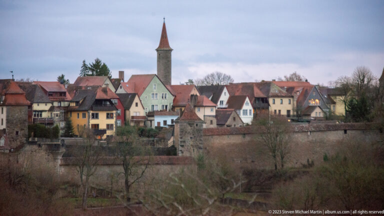 Exterior view of Rothenburg’s stone city wall, clustered pastel houses, red roofs, and a slender tower above a winter-bare slope.