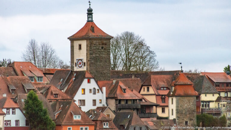 Square clock-faced tower with orange roof rising among dense red-tiled houses, with a smaller defensive tower at right.