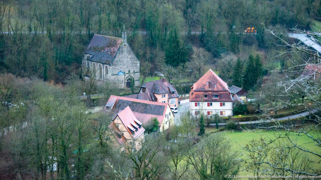 Small stone church-like building with nearby houses and red roofs in green fields and woods of the Tauber valley.