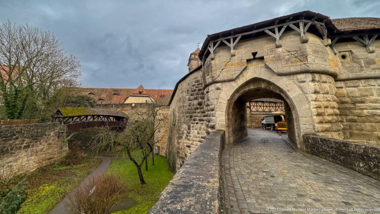 Massive stone pointed-arch gateway with covered timber wall walk, cobbled approach, and adjoining raised fortification passage.