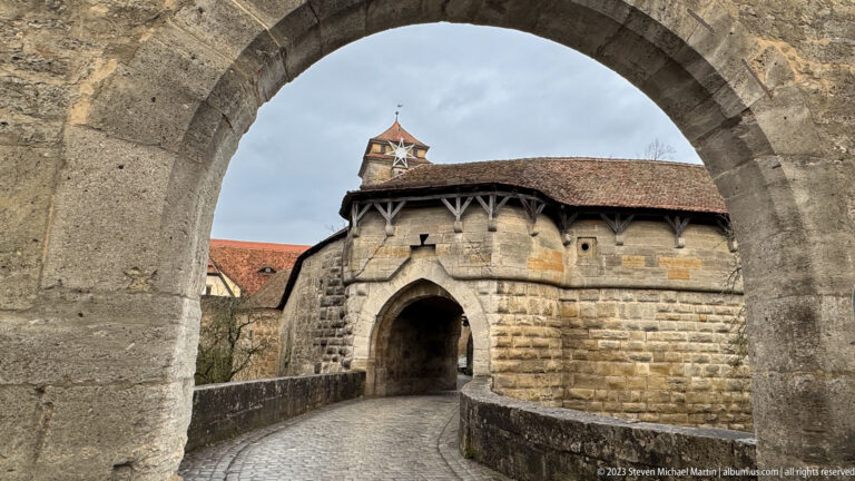 Pointed stone gateway framed through a larger foreground arch, with covered timber passage and curving cobblestone roadway.
